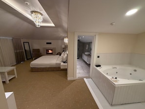 Habitación de hotel moderna con una cama grande, chimenea, baño con bañera y vista al pasillo.
