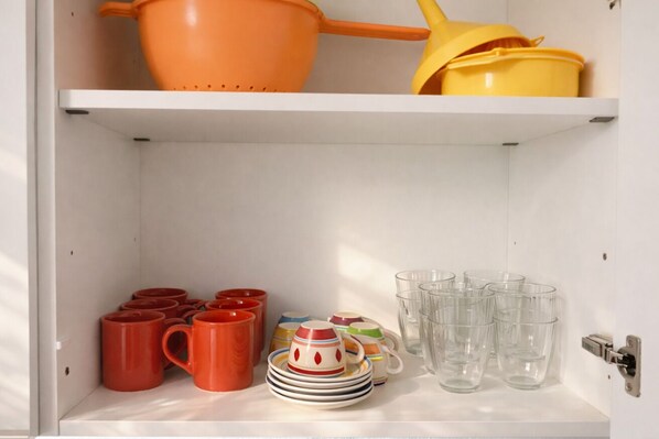 Une étagère blanche sur laquelle sont disposés divers ustensiles de cuisine, notamment des passoires orange et jaunes, des tasses rouges, des assiettes colorées et des verres transparents.