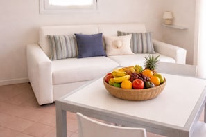 Un sofá blanco con almohadones a rayas, una mesa blanca con frutero y una lámpara sobre una estantería.