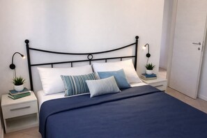 A neatly made bed with a blue bedspread, white pillows, and striped pillows. There are bedside tables with lamps and a small plant on each.