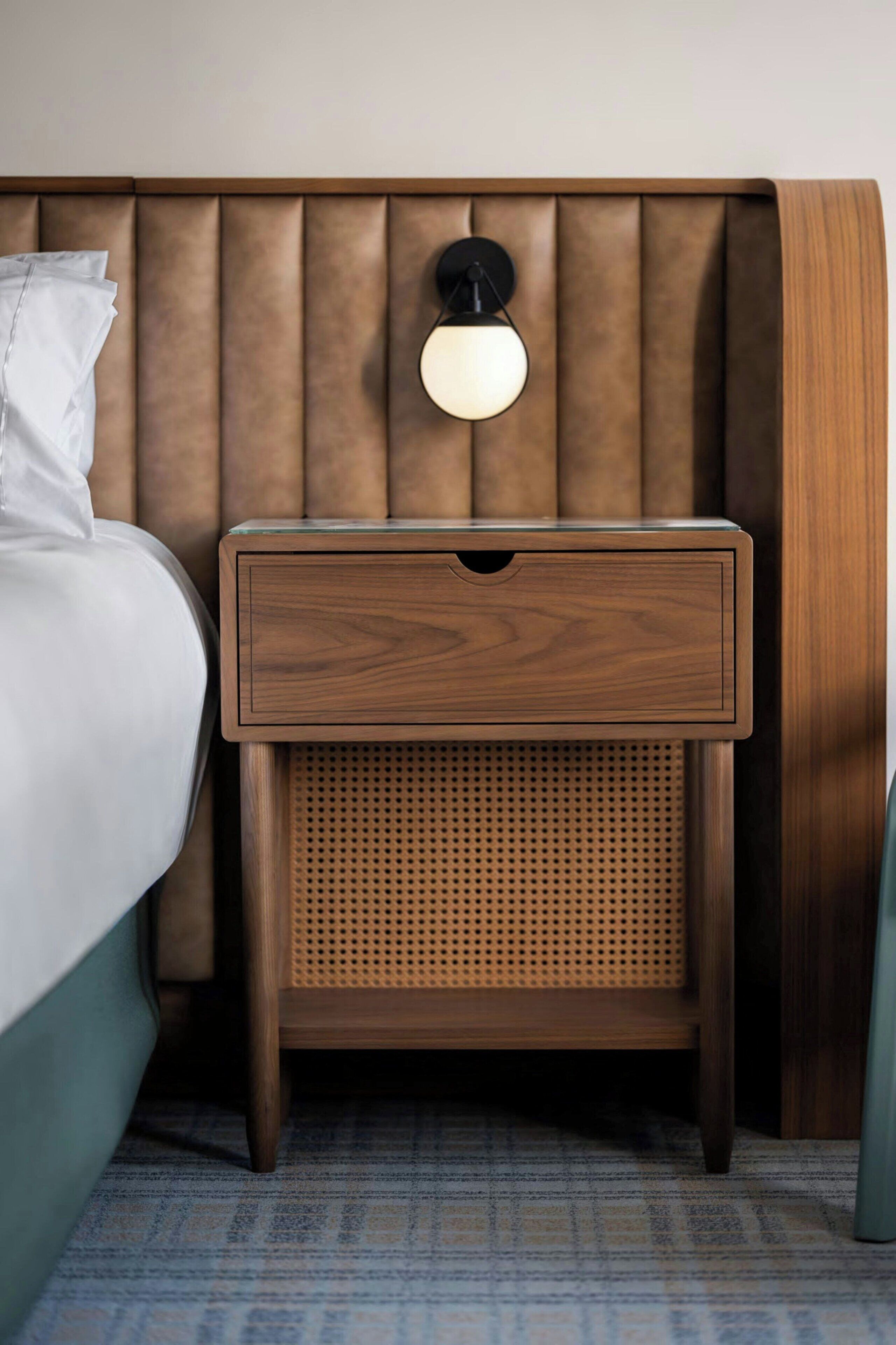 Una mesita de noche de madera con un cajón y un panel perforado, ubicada junto a una cama con cabecera blanca.