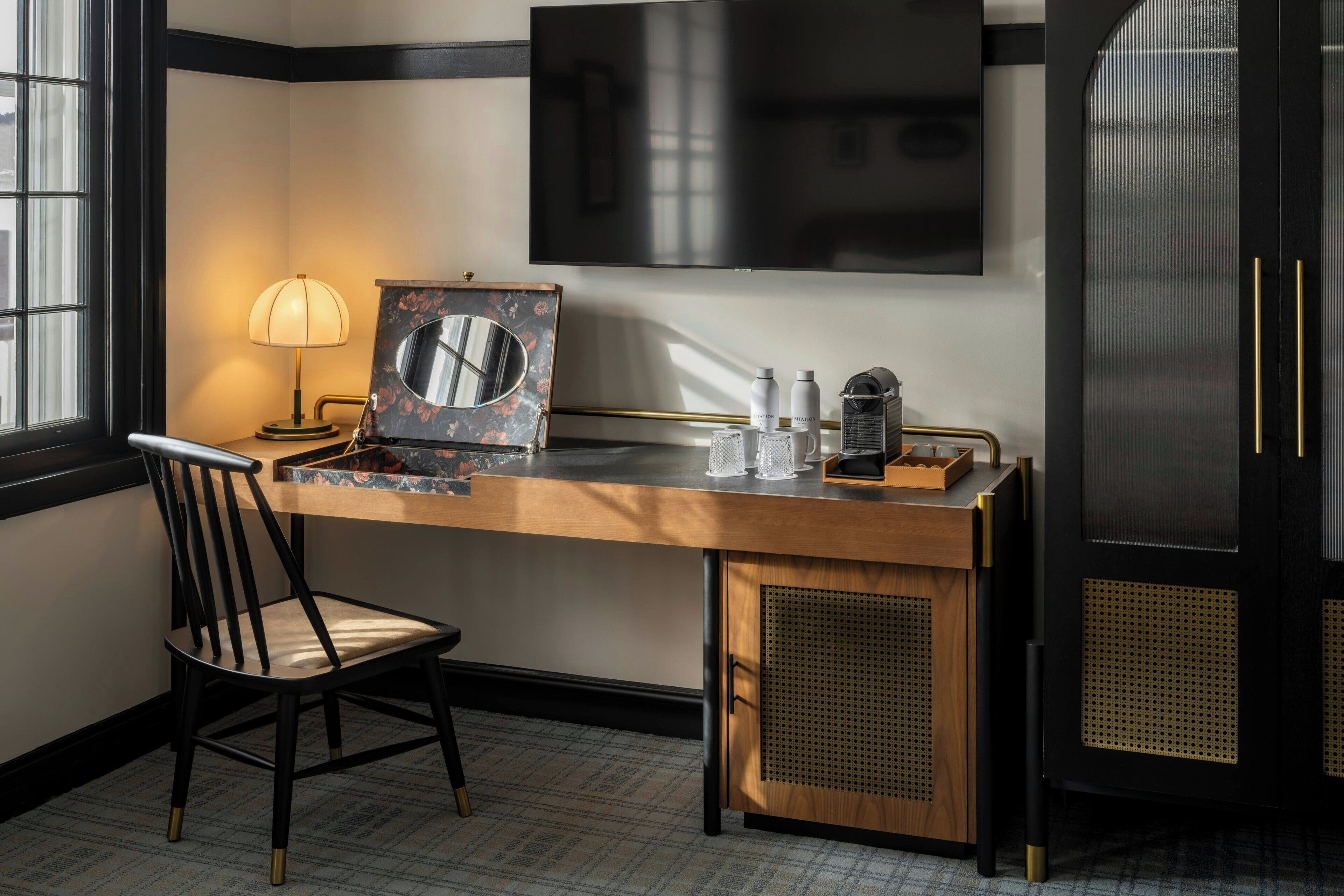 Habitación de hotel con escritorio de madera, una silla, un espejo grande, televisor de pantalla plana y un minibar.
