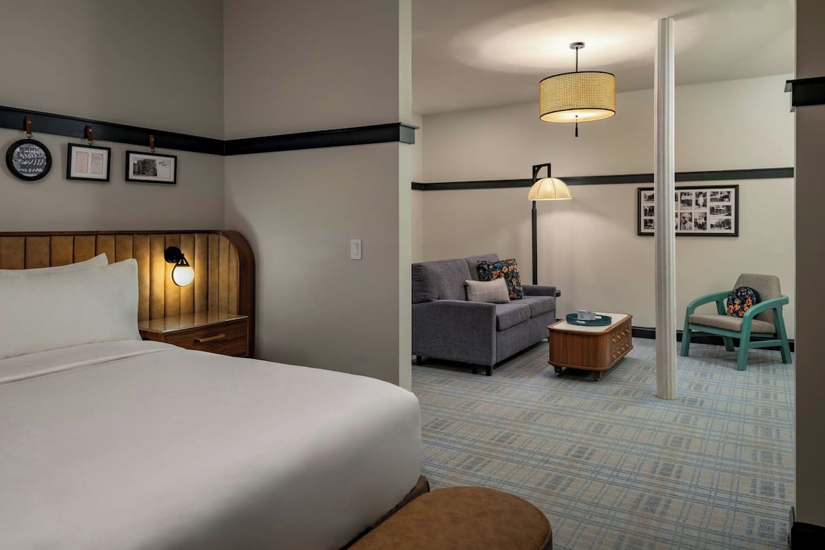 A hotel room with a bed, a nightstand, a lamp, a sofa, a chair, and a small table.