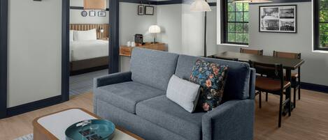 A modern hotel room with a grey sofa, a wooden coffee table, a dining area with a table and chairs, a bed with a headboard, and a large window with greenery outside.