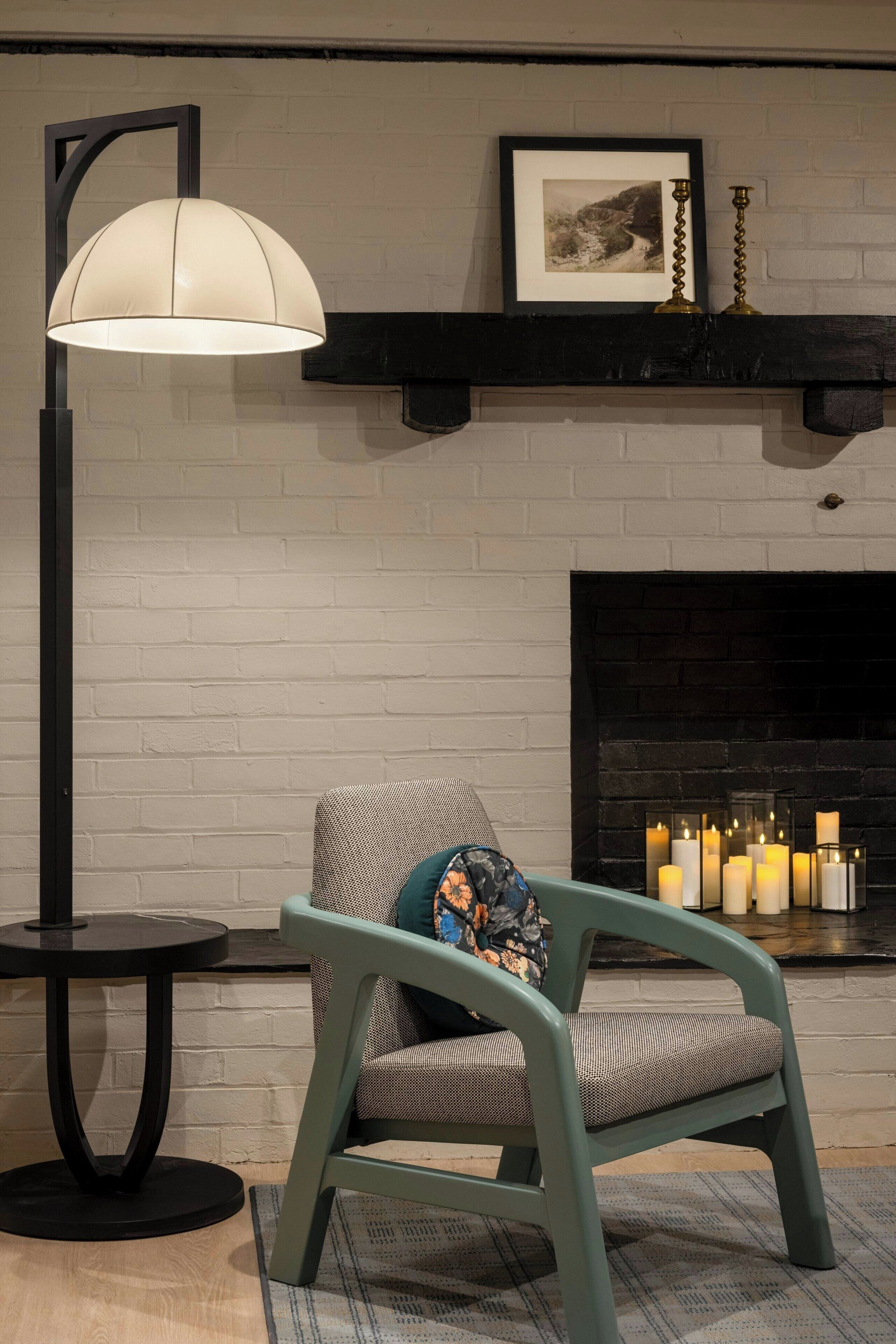Una habitación con chimenea, un cuadro colgado encima, una lámpara de pie, una mesa redonda y una silla con cojín.