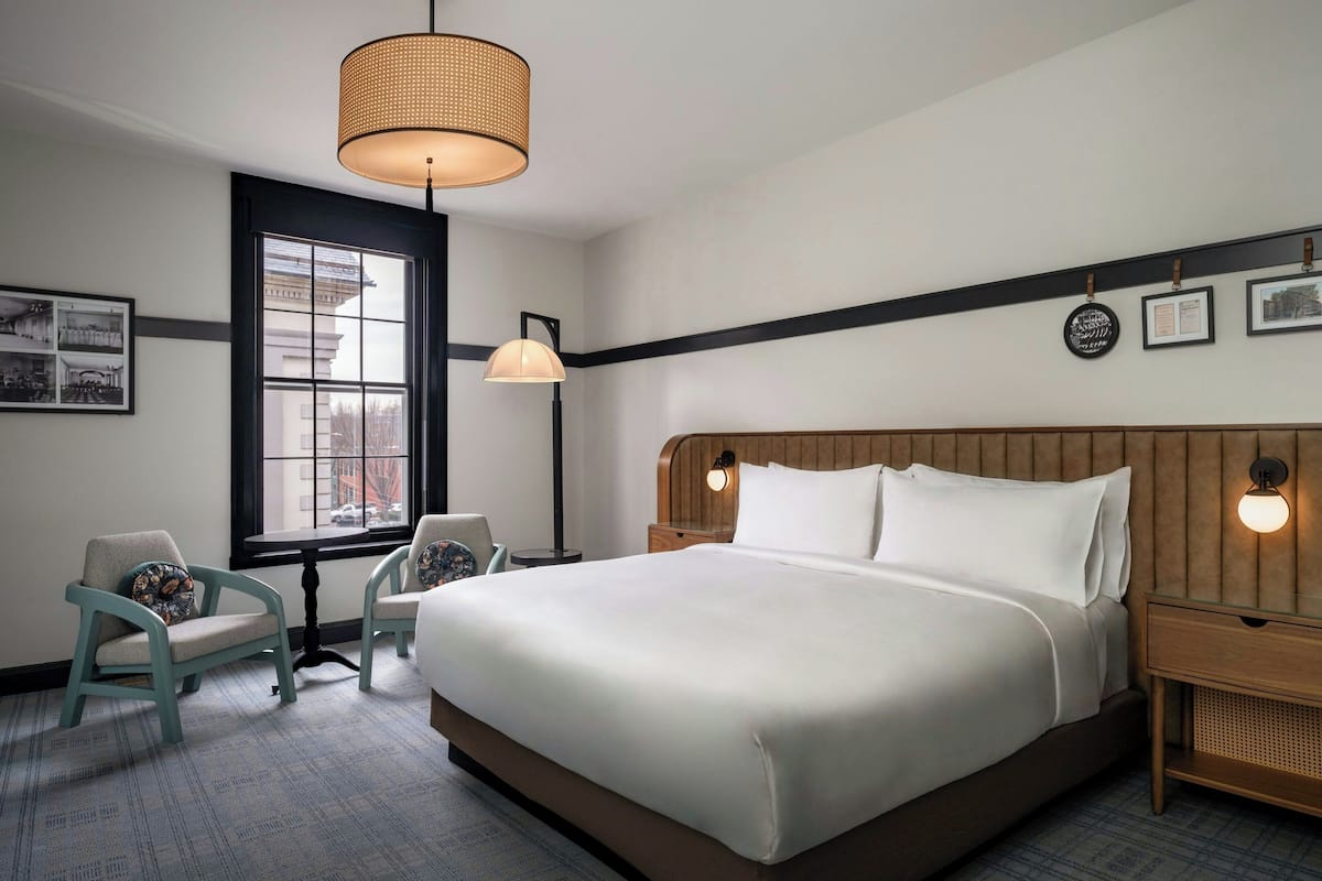A hotel room with a large bed, two chairs, a window, and wall-mounted artwork.