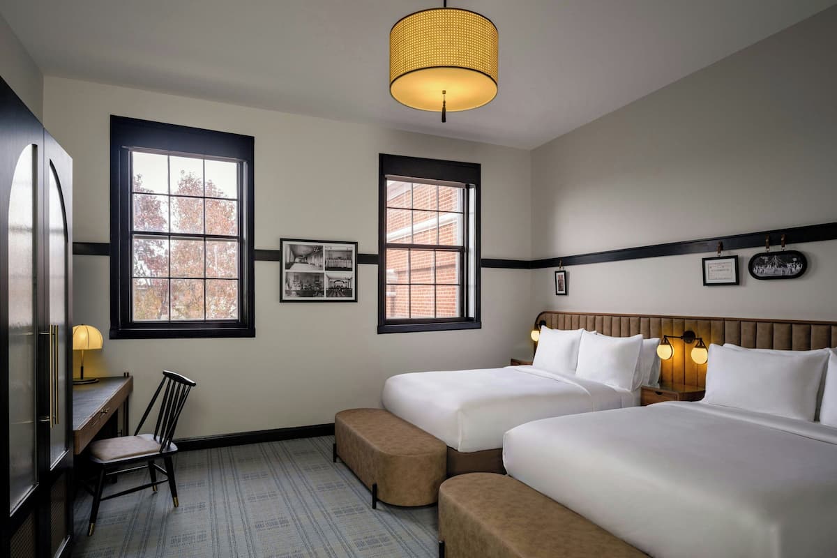 A hotel room with two beds, a desk, a chair, and two windows.