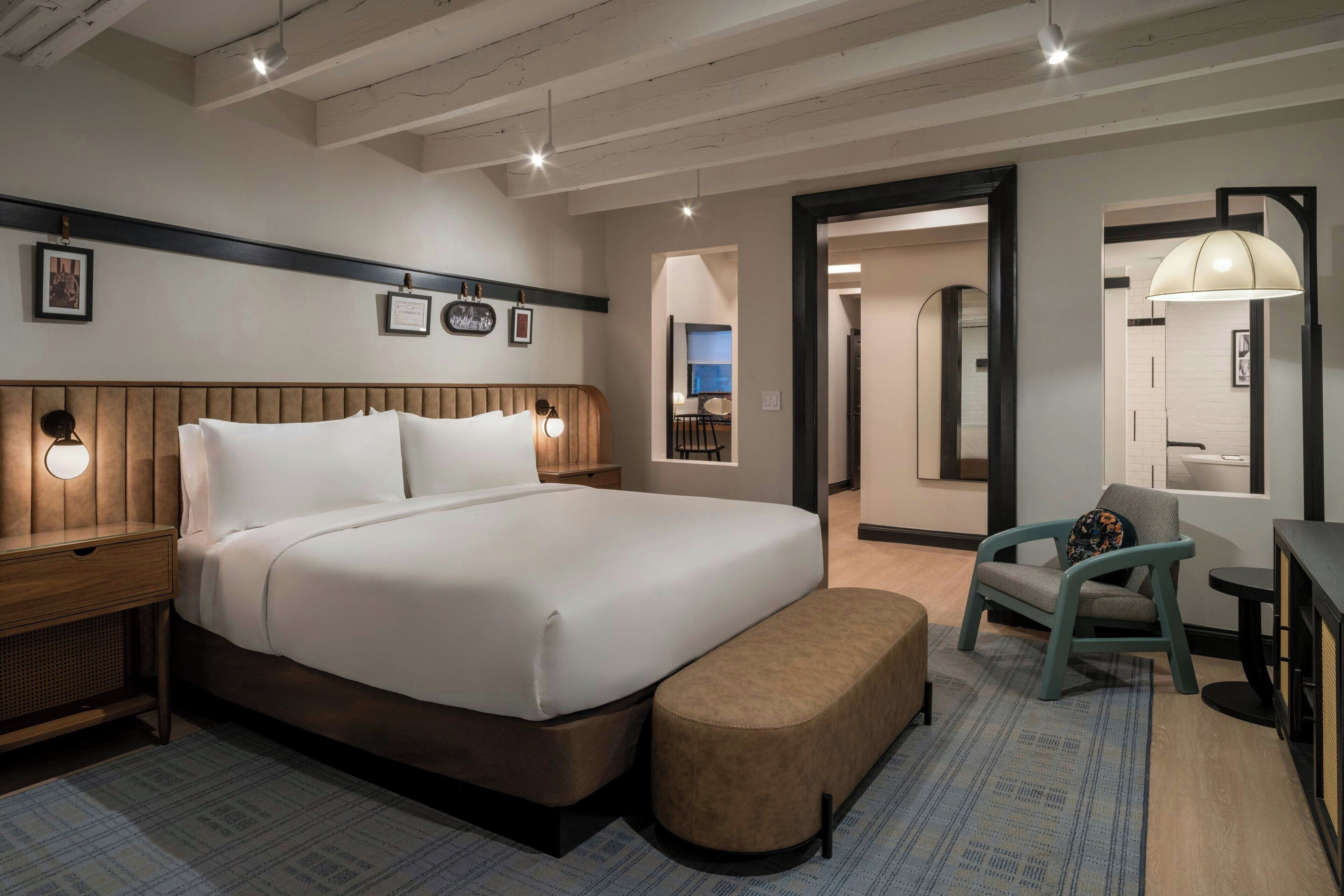 A modern hotel room with a large bed, a wooden nightstand, a bench, and a chair. There is a desk with a lamp and a mirror. The room has a wooden ceiling and walls with framed pictures on the wall.