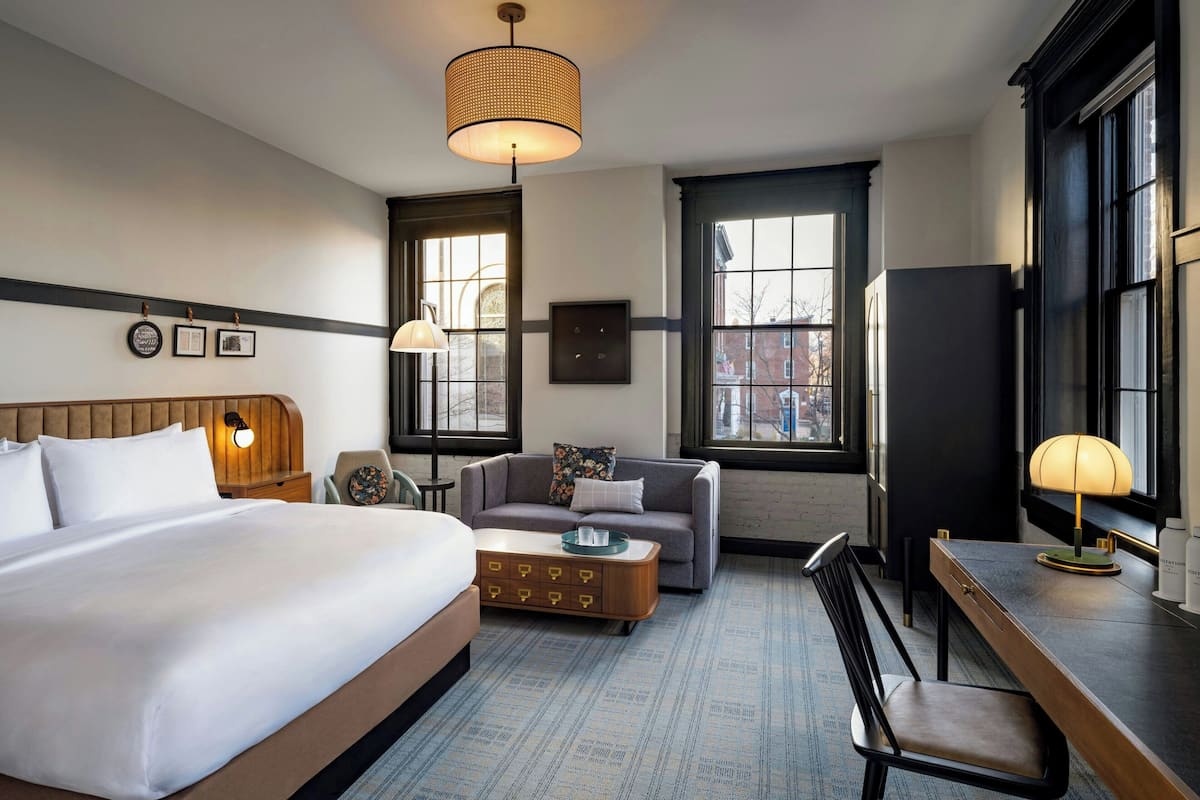 A hotel room with a large bed, a sofa, a desk, and a chair. There are two windows, a wall clock, and a lamp on the desk.