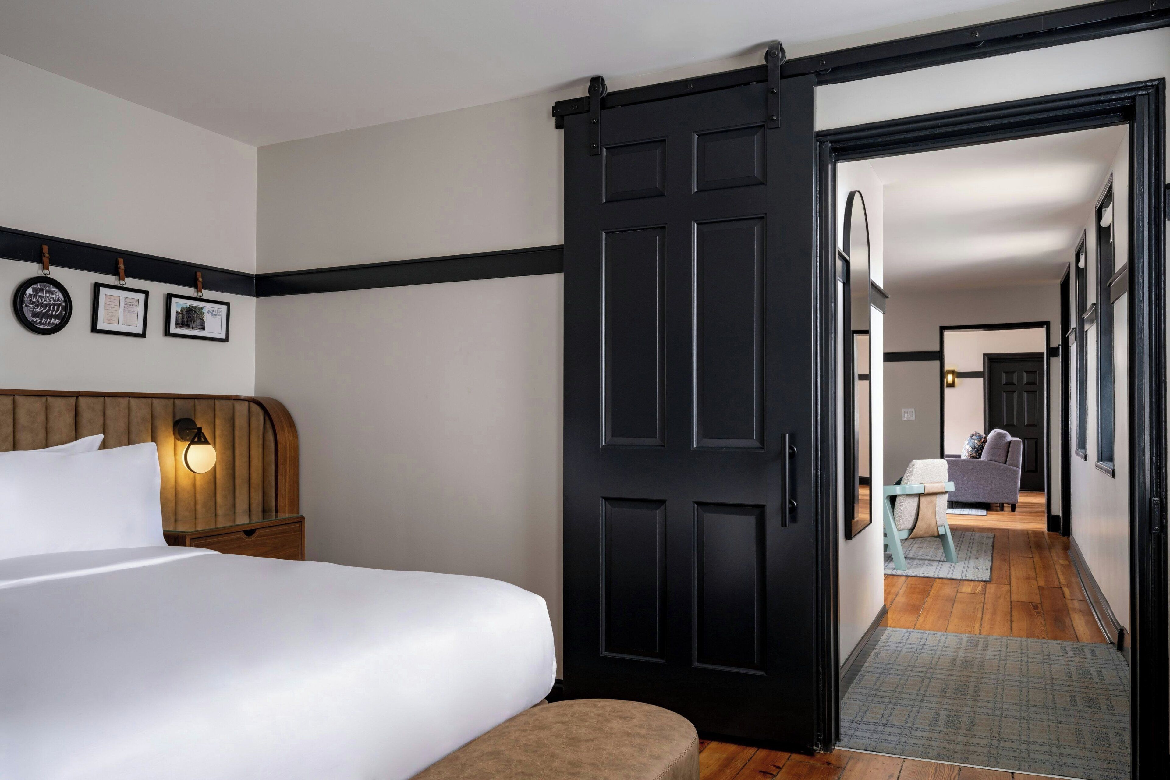 A hotel room with a bed, a nightstand, a wall-mounted light, and a sliding door leading to a hallway with a sofa and a table.