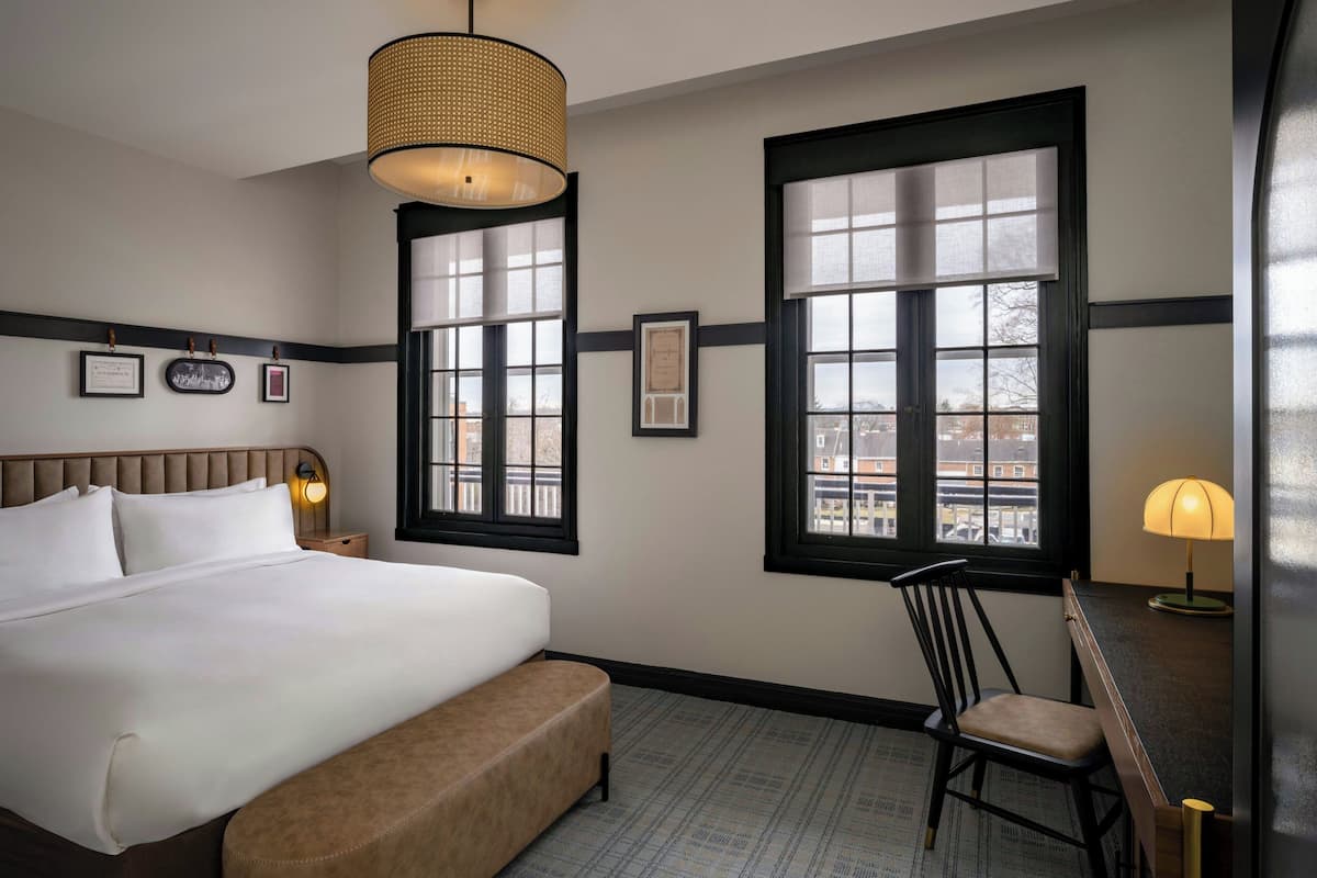 A hotel room with a large bed, a chair, a desk, and two windows with blinds.