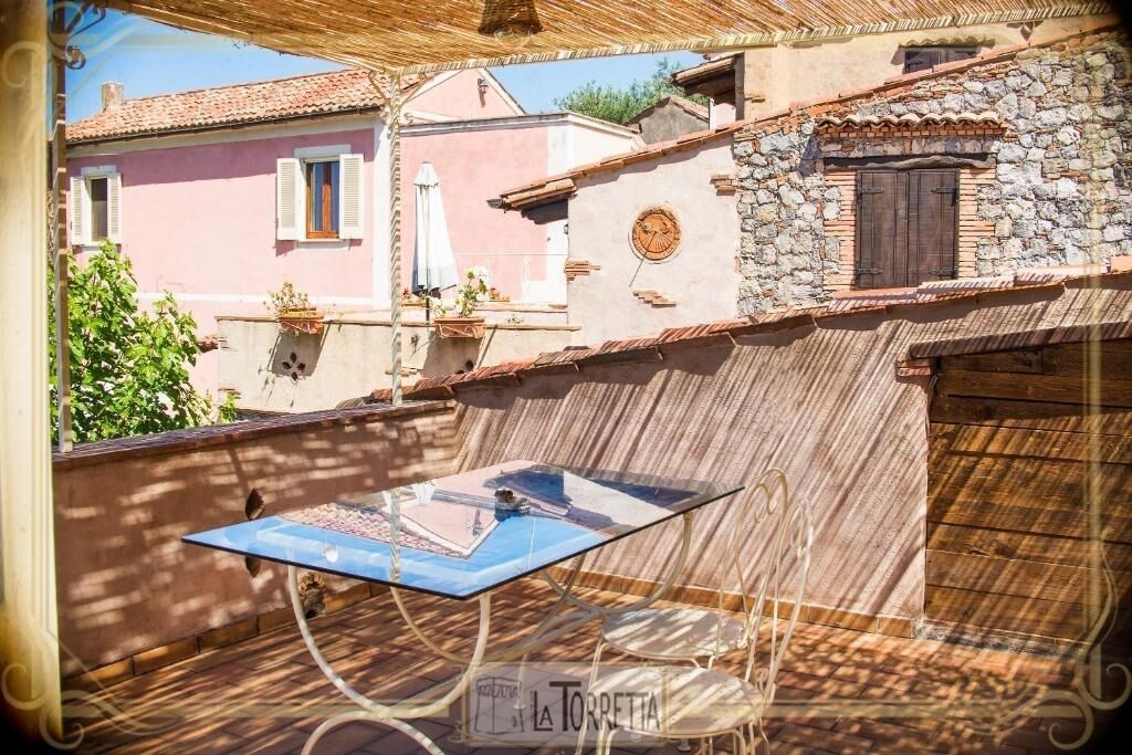 A terrace with a glass table and chairs, overlooking a pink building with a stone wall and a wooden door.