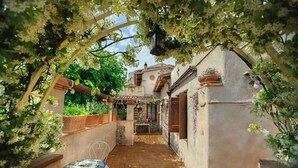 A narrow courtyard with a tiled floor, a small table with a chair, and a pergola covered with climbing plants.