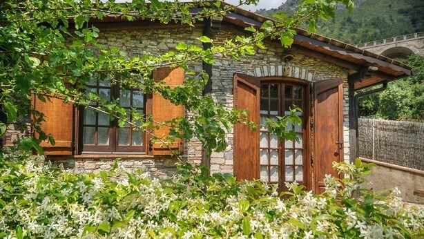 A stone house with wooden shutters, a wooden door, and a stone bridge in the background.