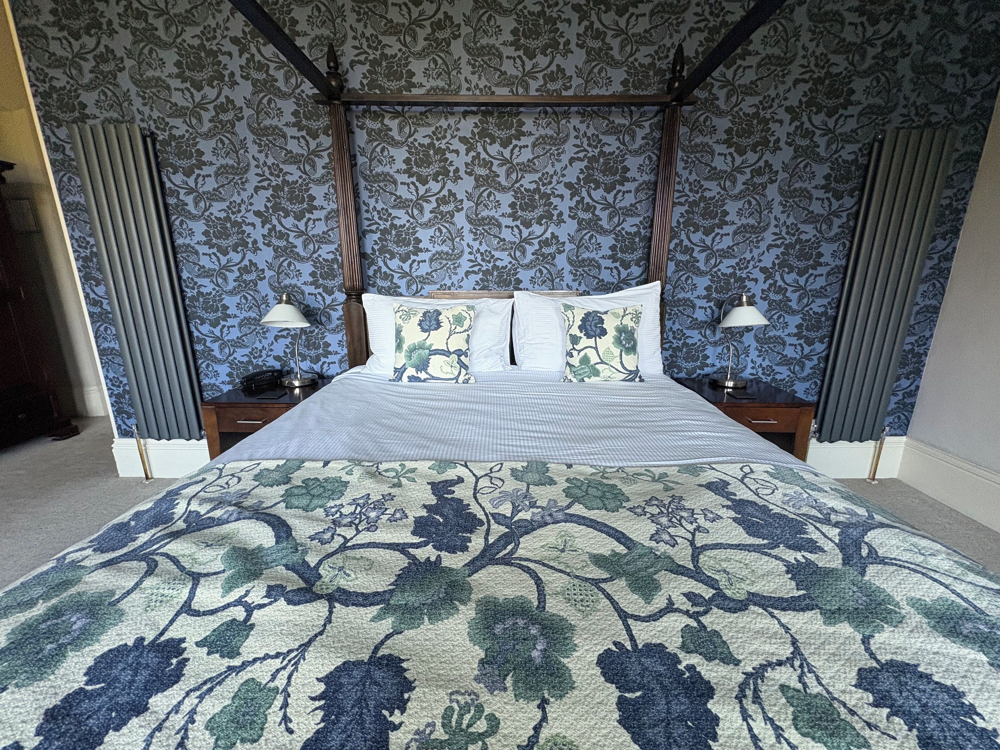 A four-poster bed with a floral-patterned bedspread, two bedside tables with lamps, and a patterned wallpaper background.
