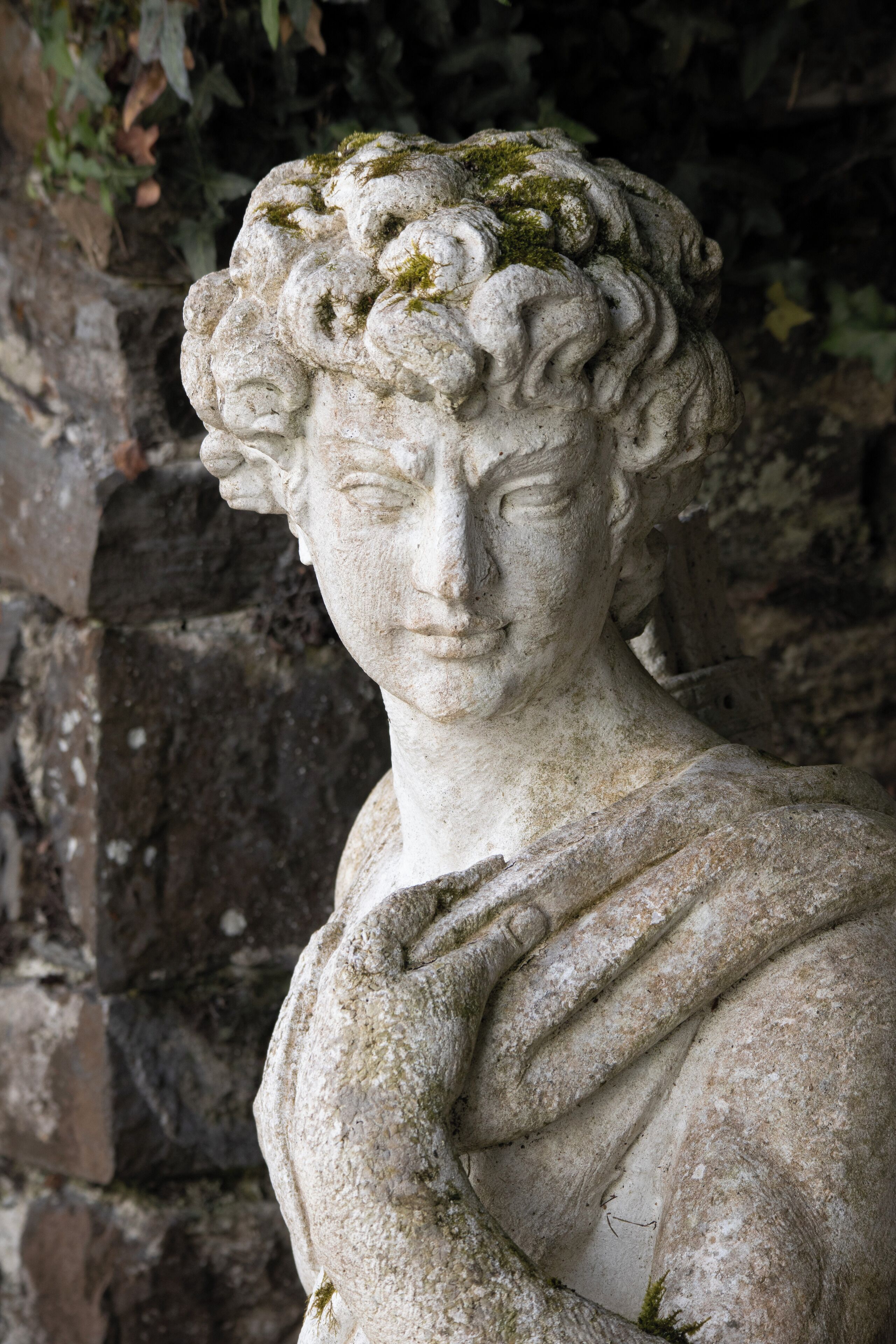 Una escultura de piedra de una figura clásica con cabello rizado y una túnica, frente a un muro de piedra.