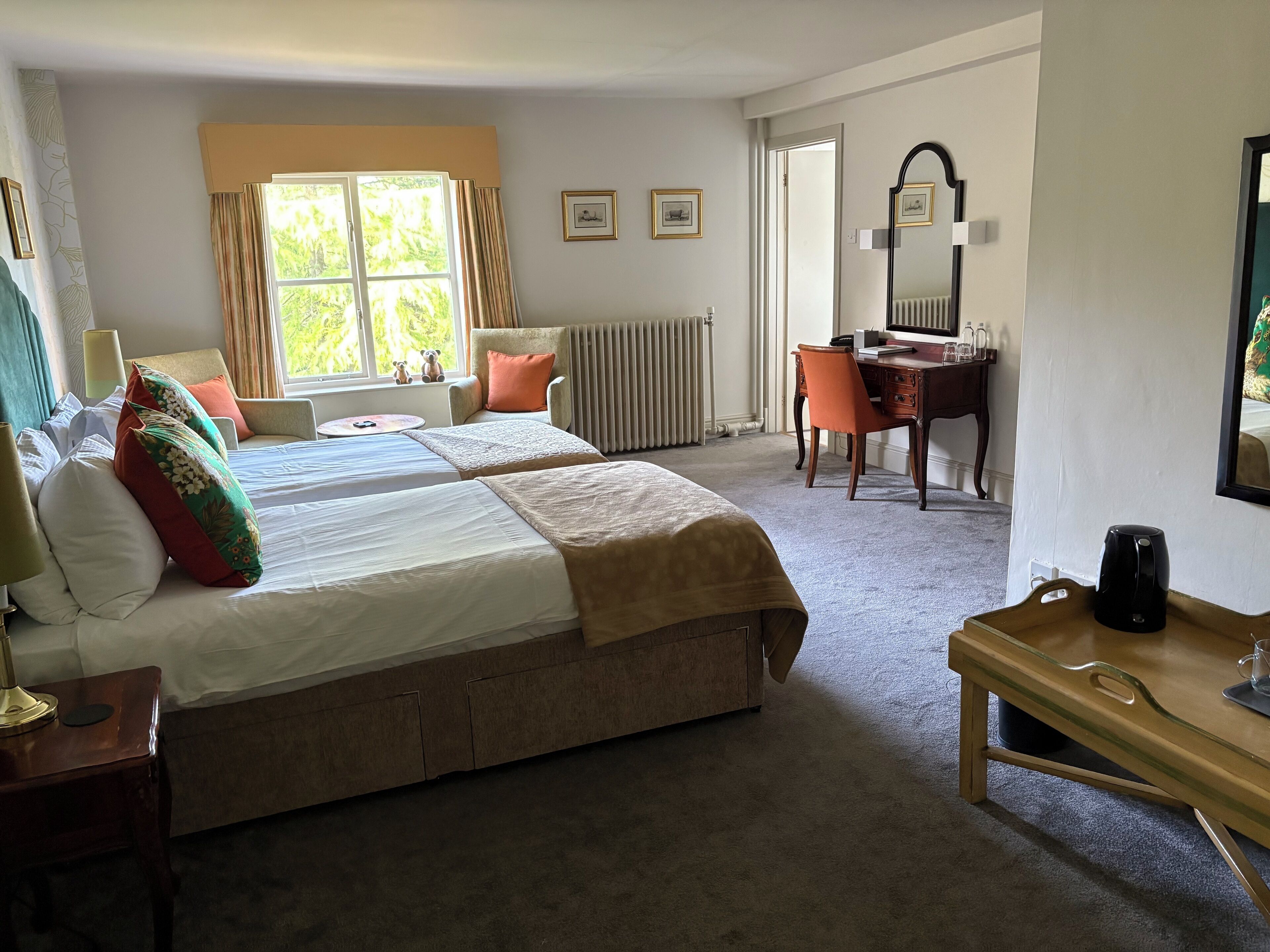 A hotel room with two beds, a desk, a chair, a radiator, and a window with curtains.