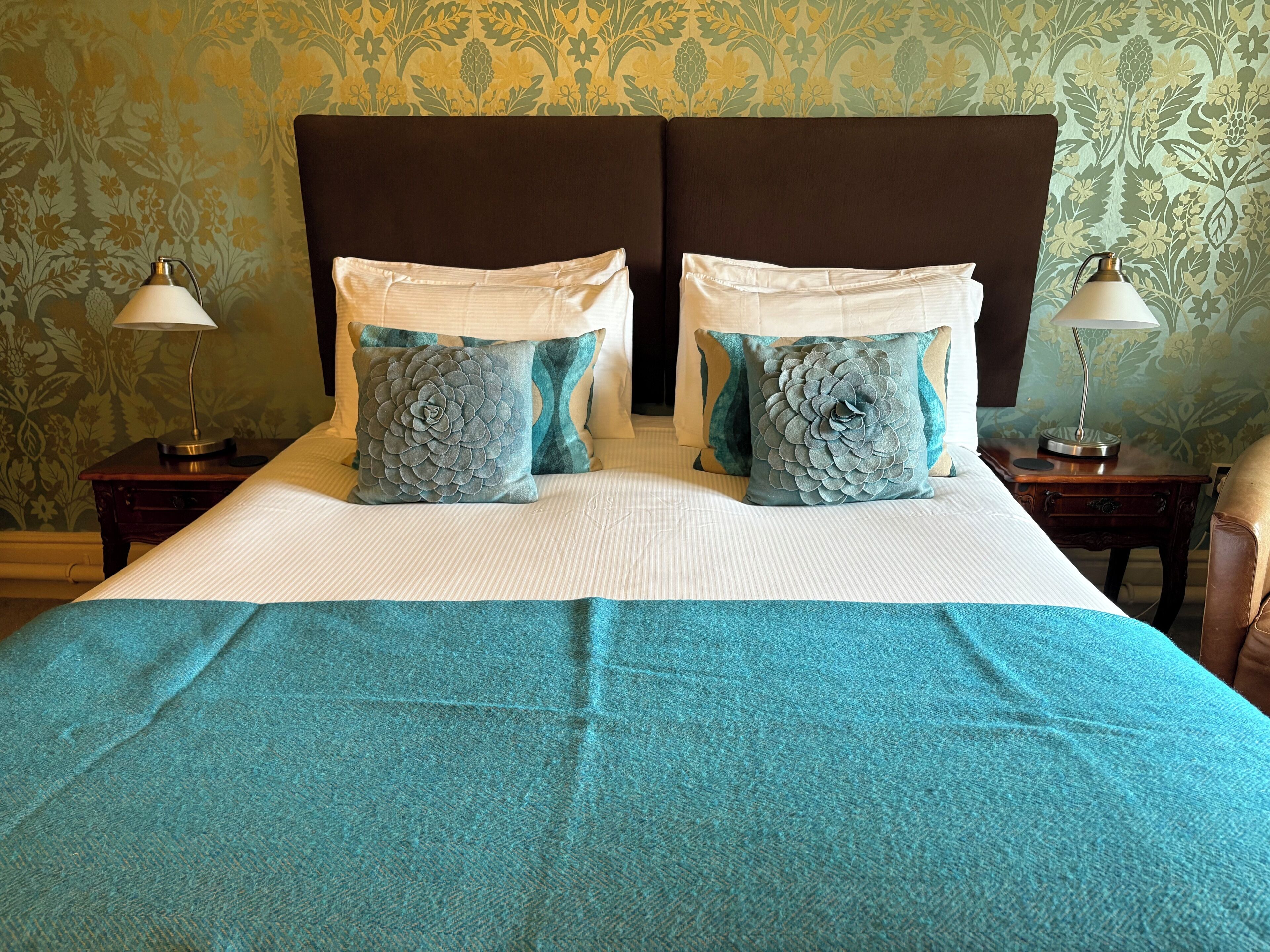 Una cama bien hecha con un edredón color aguamarina, dos almohadas decorativas y dos lámparas de noche sobre mesitas de madera.