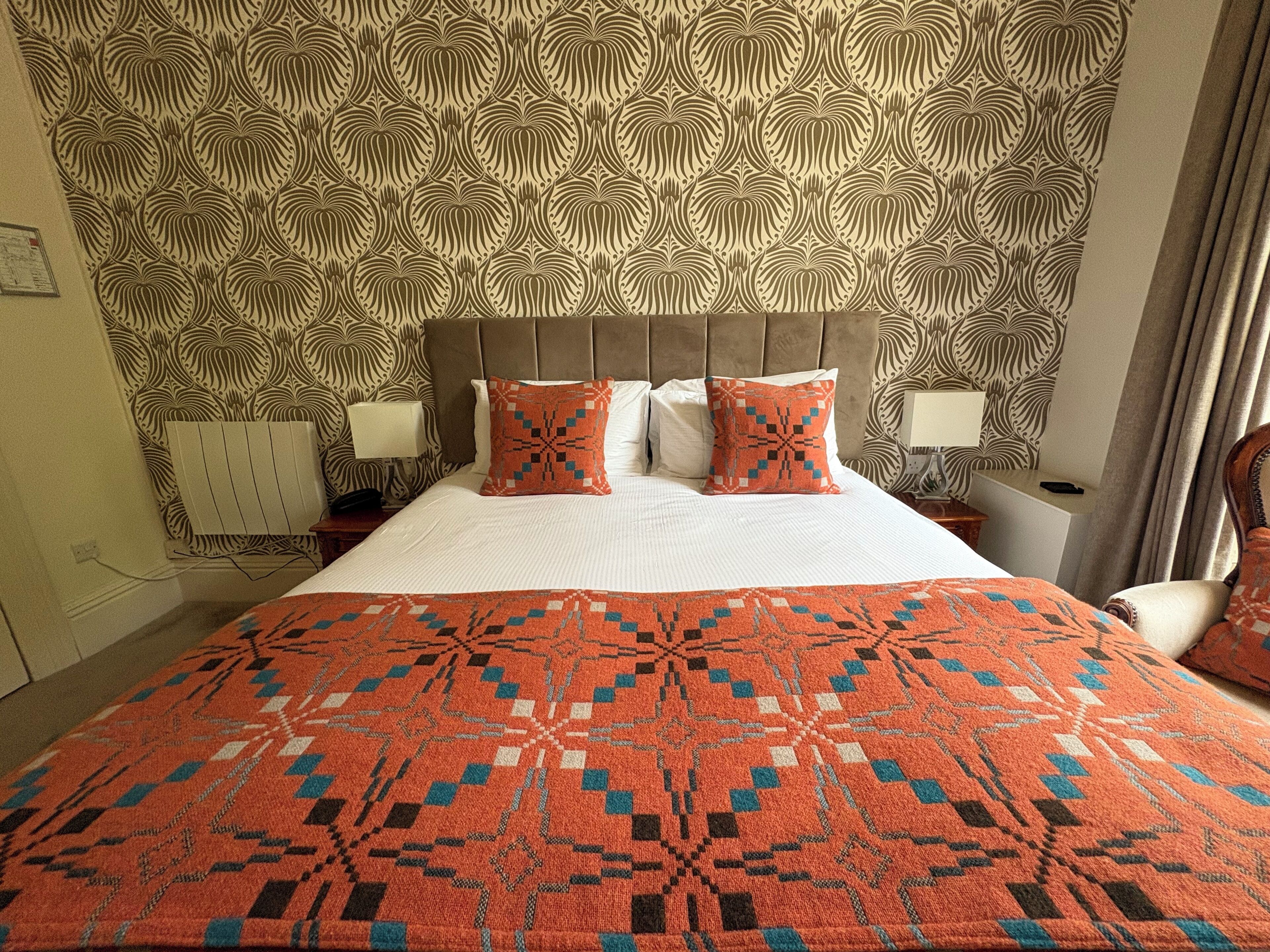 A hotel room with a patterned headboard, two pillows with geometric designs, a red and blue patterned bedspread, and a nightstand with a lamp.