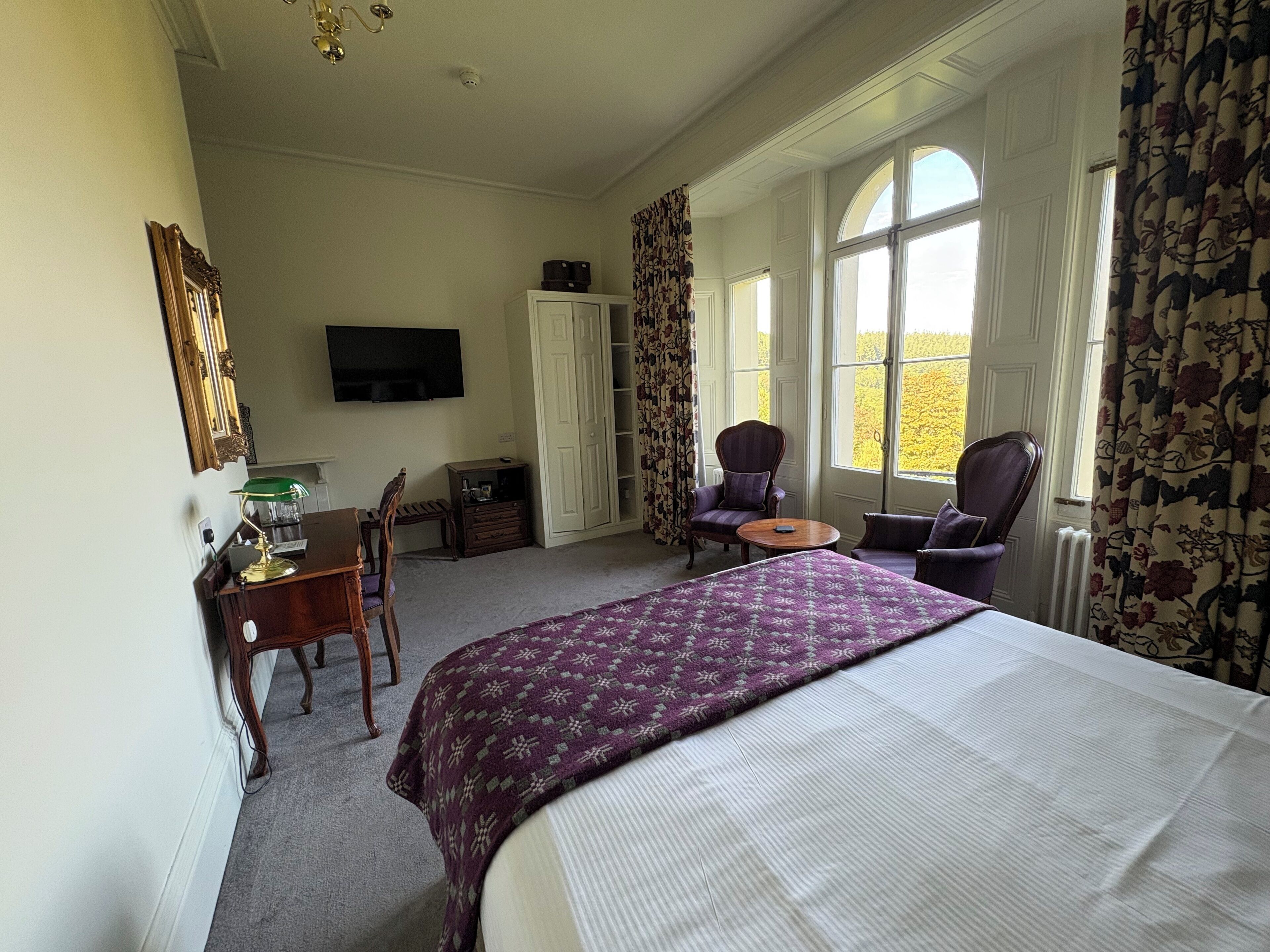 A hotel room with a bed, a desk with a chair, a television, and a window with curtains.