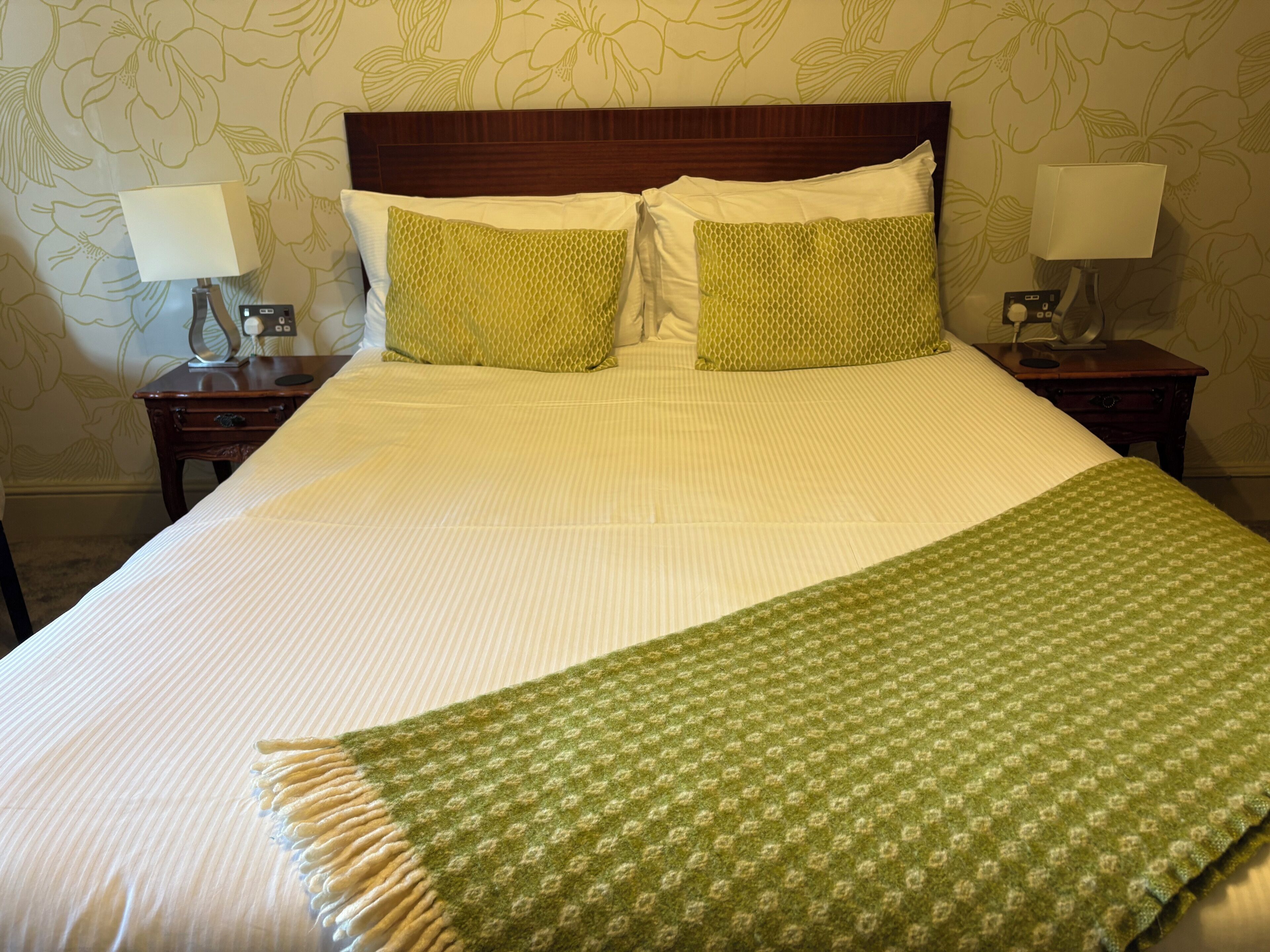 A neatly made bed with a green patterned throw, two bedside tables with lamps, and floral wallpaper.