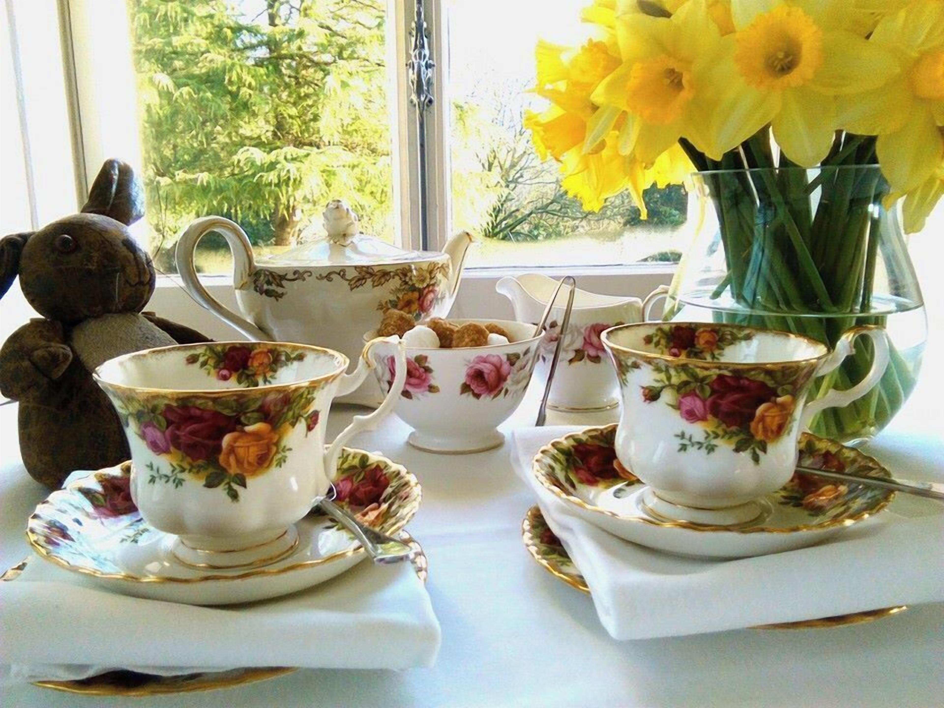 A table set with floral-patterned tea cups, saucers, and a teapot, with a vase of yellow flowers and a teddy bear on the windowsill.