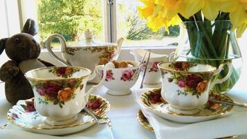 A table set with floral-patterned tea cups, saucers, and a teapot, with a vase of yellow flowers and a teddy bear on the windowsill.