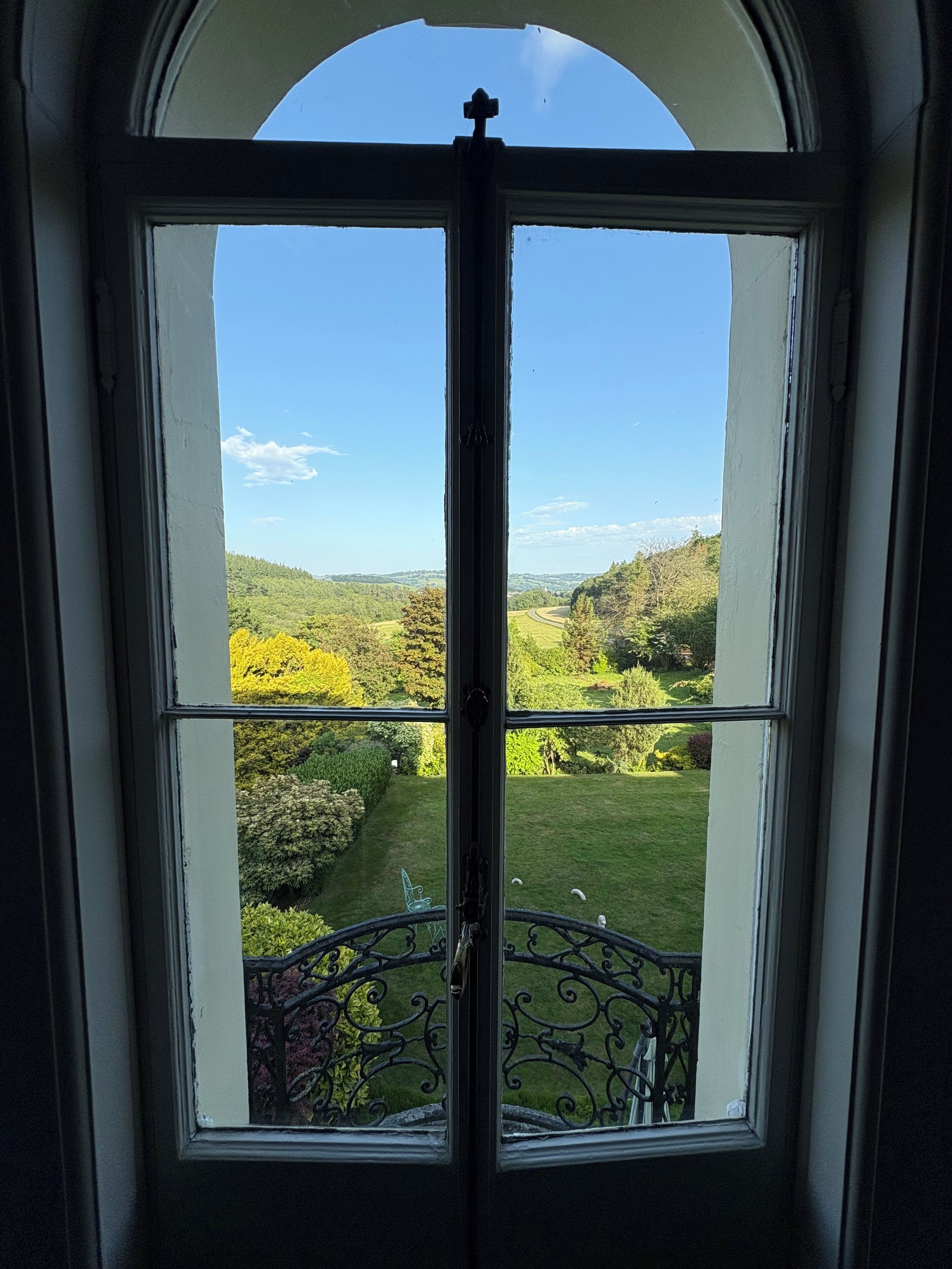 Une vue à travers une fenêtre ornée d’une rampe en fer forgé, donnant sur un paysage verdoyant avec des arbres et un ciel dégagé.