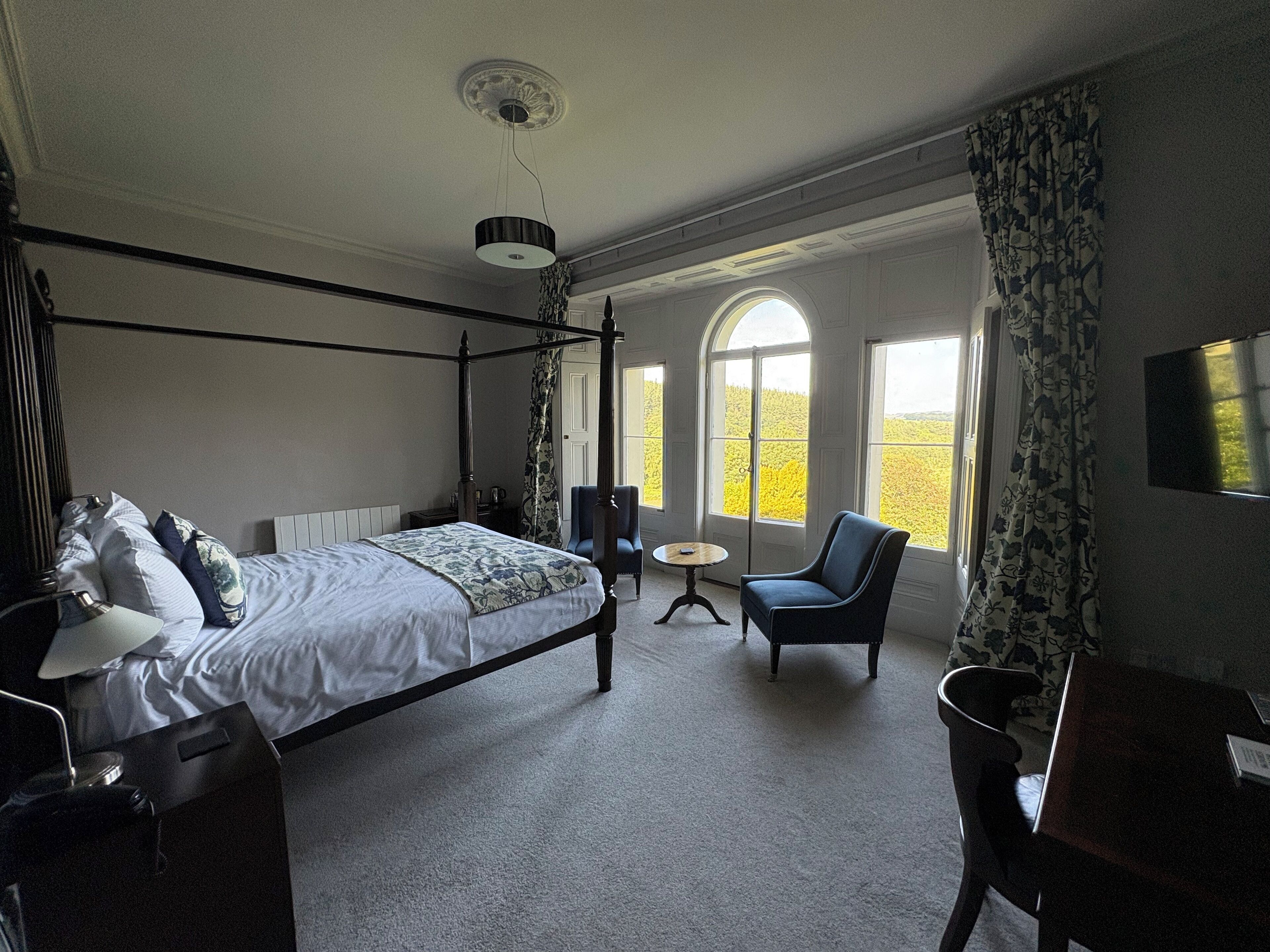 A four-poster bed with a canopy, a small round table, a chair, and a desk with a flat-screen TV.