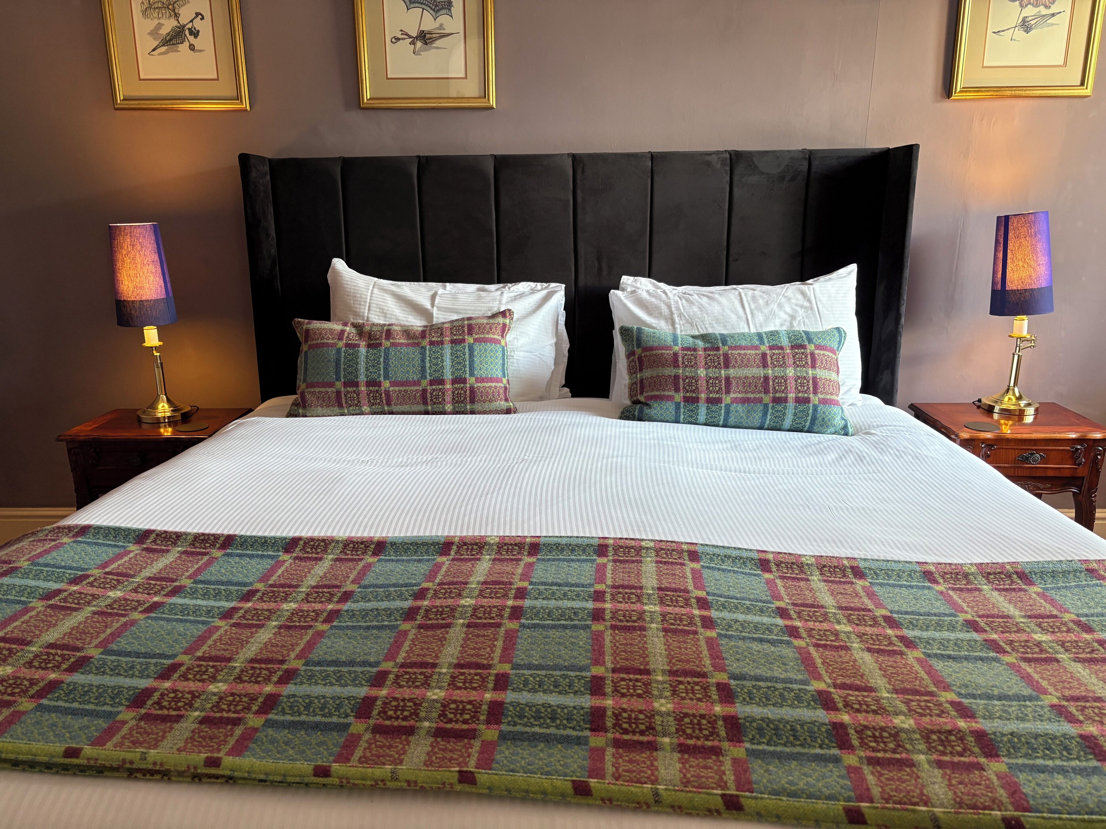 Una cama bien hecha con una colcha escocesa, dos almohadas a juego y dos lámparas de noche sobre mesitas de madera.