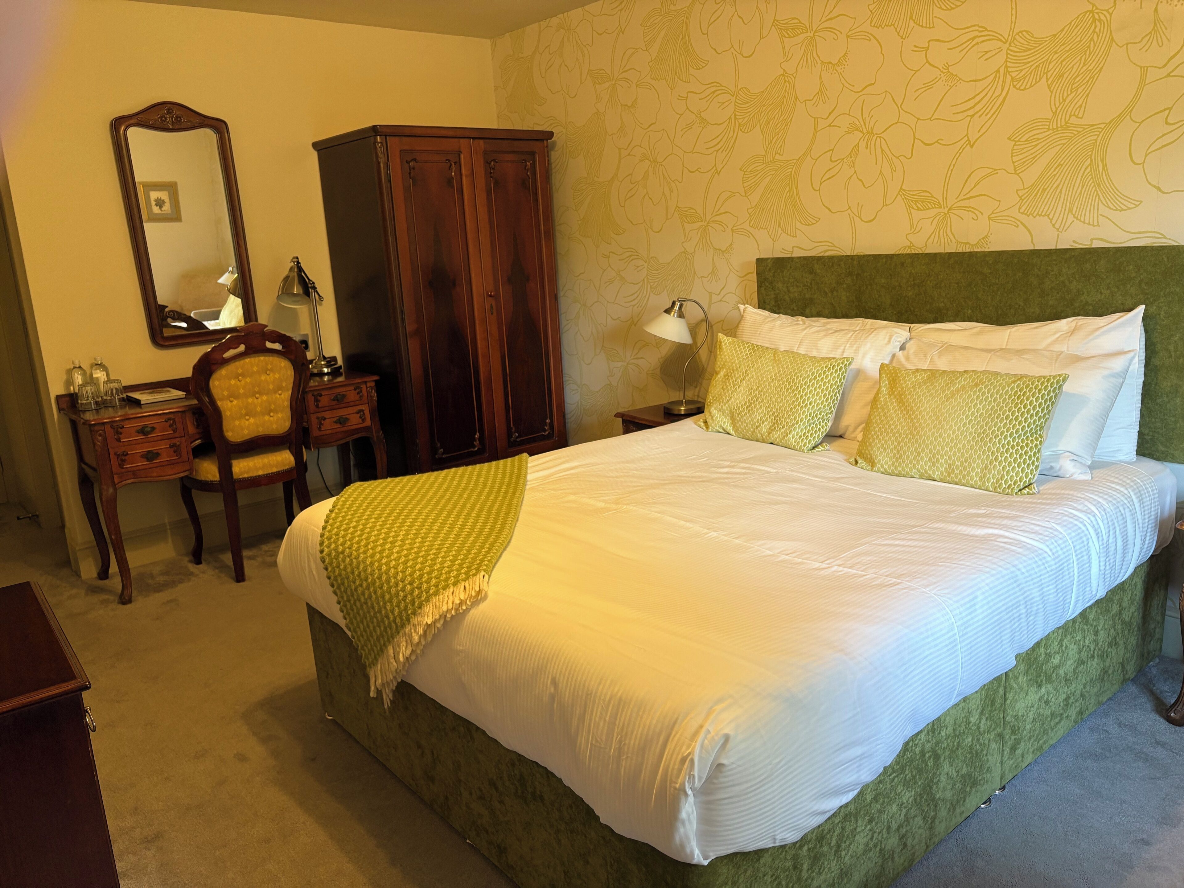 A neatly made bed with a green headboard, a wooden wardrobe, a desk with a chair, and a mirror on the wall.