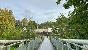 A pedestrian bridge with metal railings, a wet pathway, and buildings in the background. - Batheaston Terrace (Batheaston)
