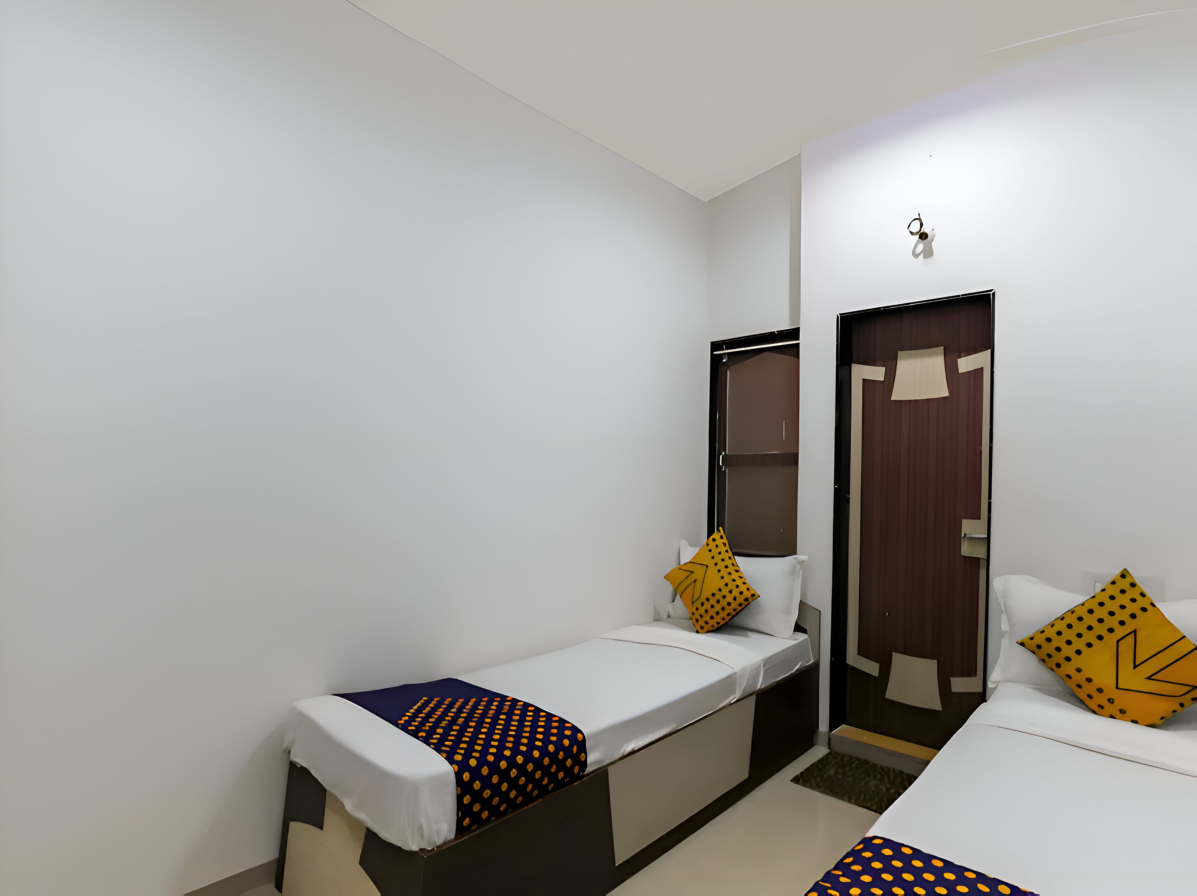 A small, clean room with two single beds, each with a white mattress and a patterned blanket. There is a wooden door and a mirror on the wall.