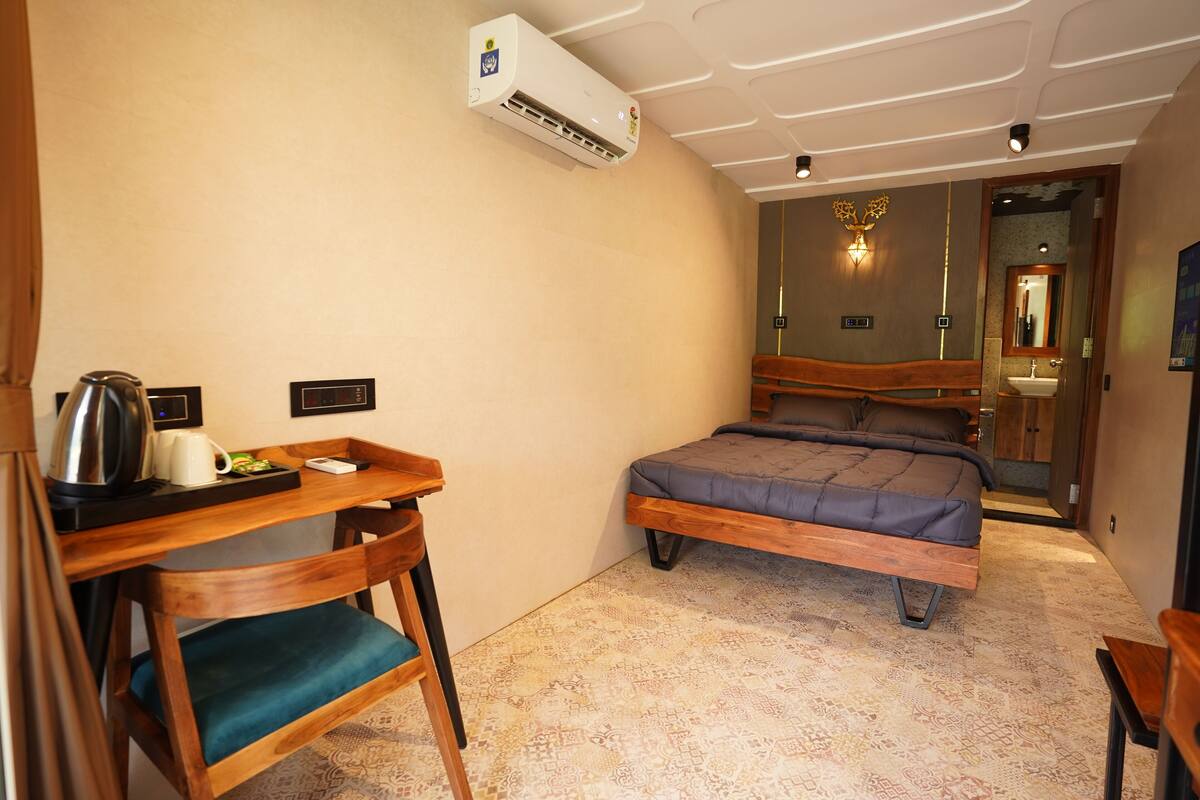 A compact hotel room with a bed, a desk with a kettle, a chair, and a small table.