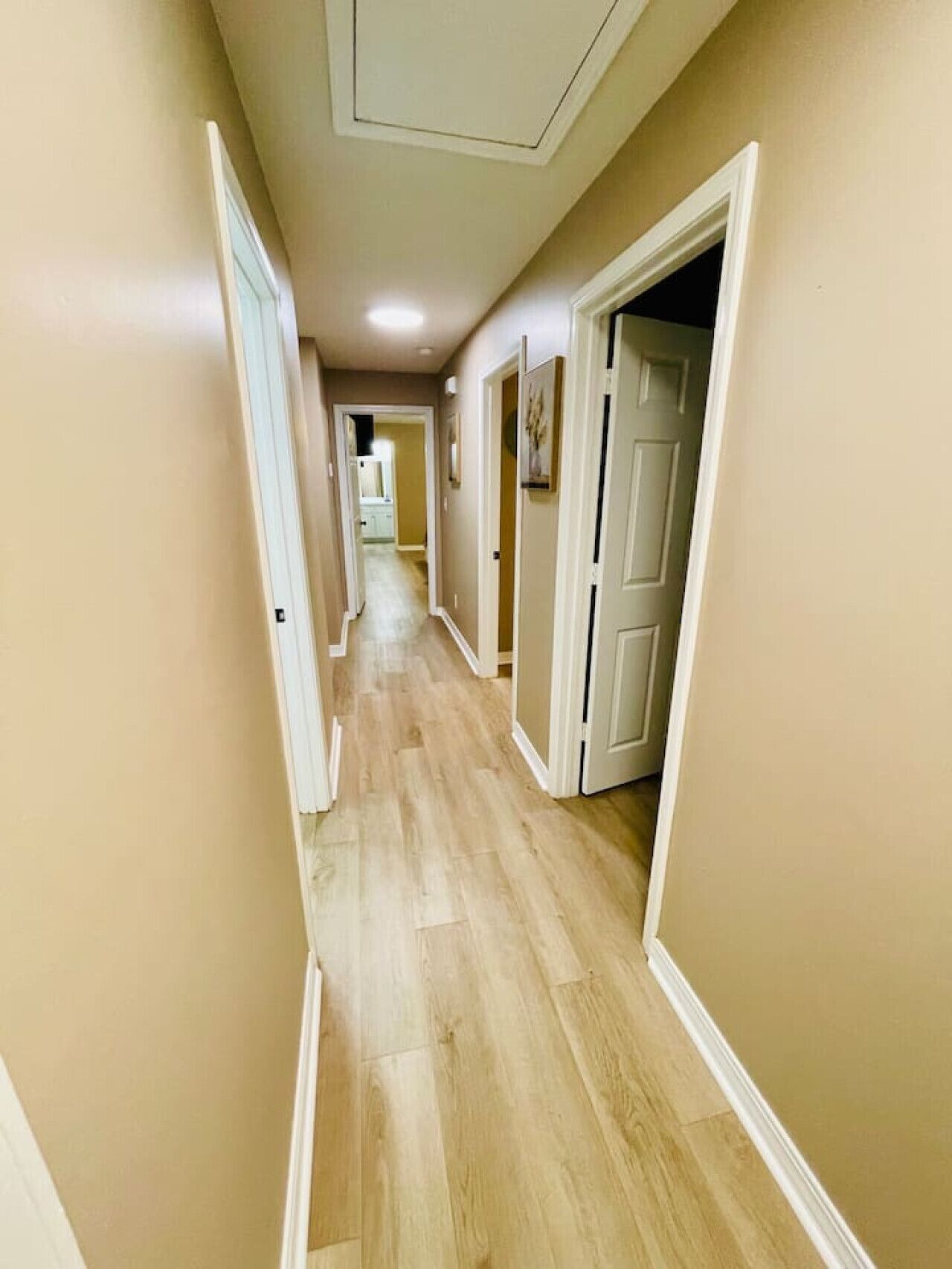 A hallway with beige walls and light wooden flooring, leading to a room with a door at the end.