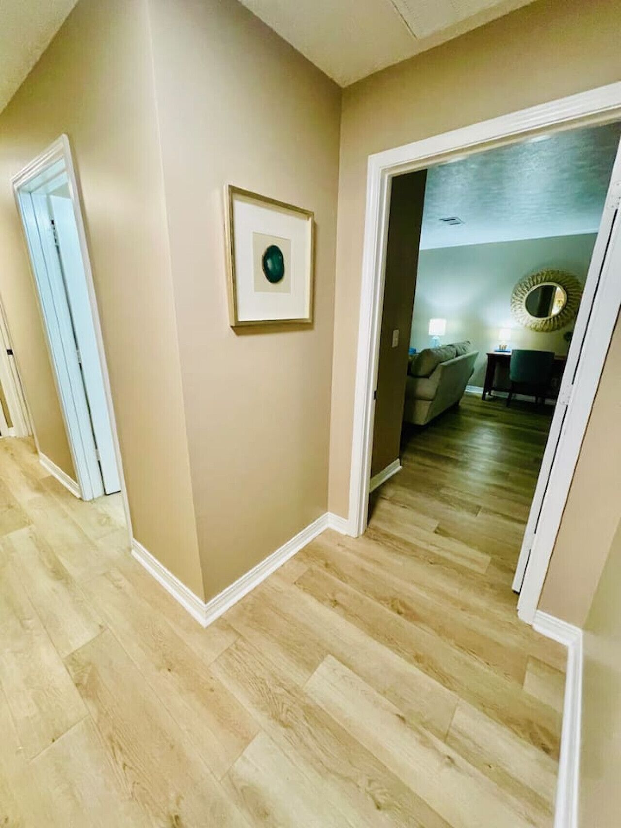 Een gang met behang in beige tinten en een houten vloer, die leidt naar een kamer met een stoel en een ronde spiegel aan de muur.
