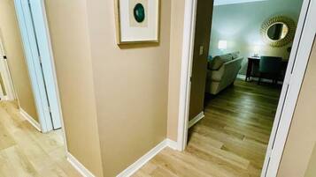 Een gang met behang in beige tinten en een houten vloer, die leidt naar een kamer met een stoel en een ronde spiegel aan de muur.