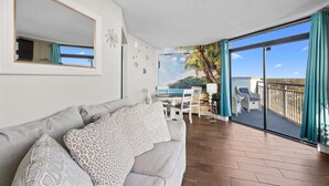 A modern living room with a sofa, a dining table with chairs, and a balcony with a view of the sea.