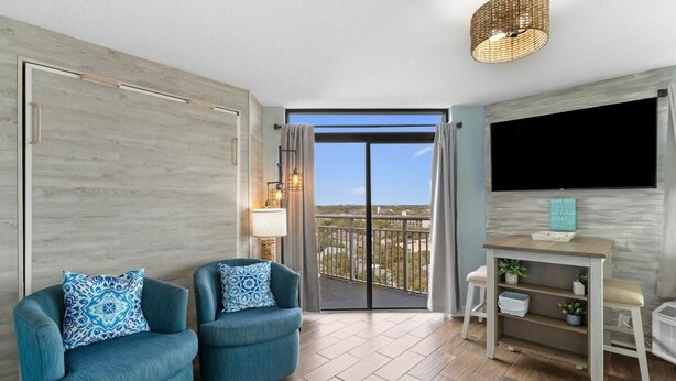 A modern living room with two blue armchairs, a wooden floor, a TV mounted on the wall, and a balcony with a view. - Ocean View Retreat: Includes Breakfast 14th Flr (Myrtle Beach)