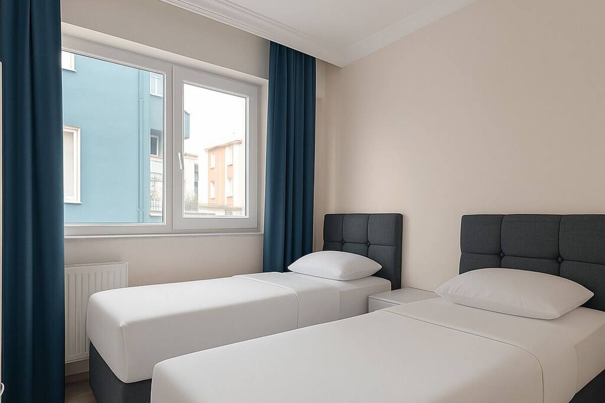 Una habitación con dos camas individuales, una ventana con cortinas azules y vistas a edificios.