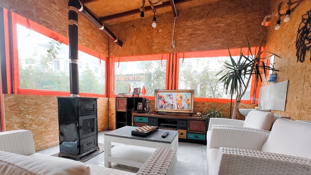A cozy living room with a fireplace, white wicker furniture, and a large window with orange curtains. - Paradise Pansiyon Otel (Bursa)