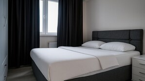 A modern bedroom with a grey upholstered bed, white linens, a grey headboard, a bedside table with drawers, and a window with dark curtains. - Paradise Pansiyon Otel (Bursa)
