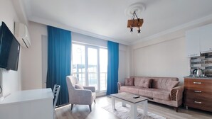 A modern living room with a sofa, armchair, and coffee table. There is a TV mounted on the wall, a kitchen area with a stove, and a balcony with blue curtains. - Paradise Pansiyon Otel (Bursa)