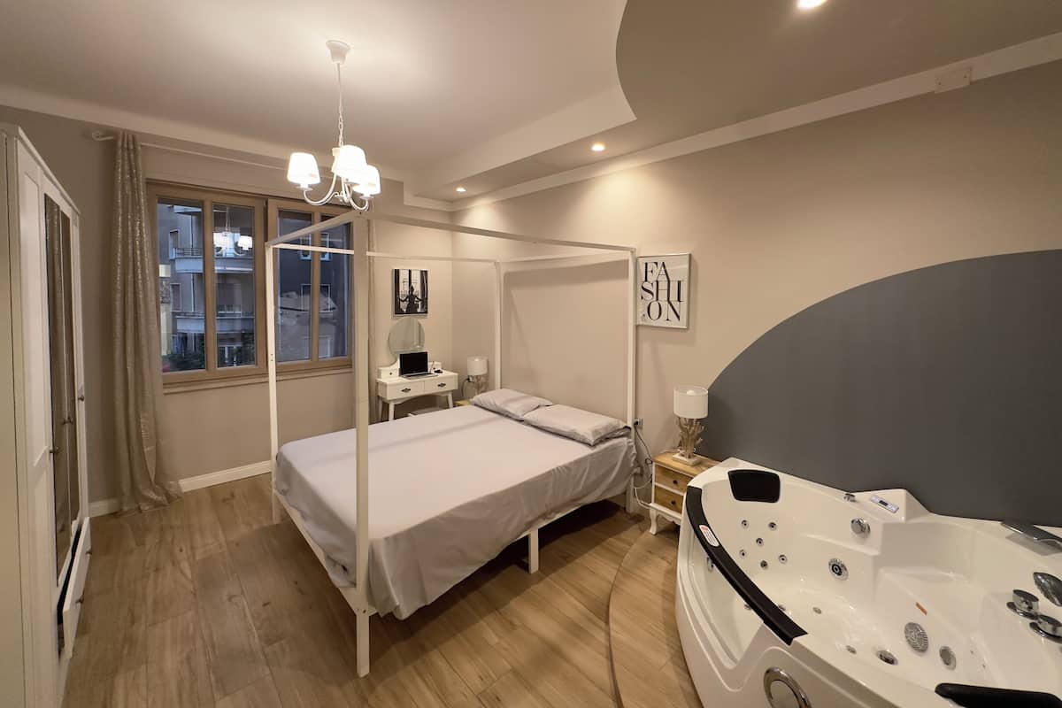 Un dormitorio con una cama con dosel, una mesita de noche con lámpara, un ventanal grande y una bañera de hidromasaje.