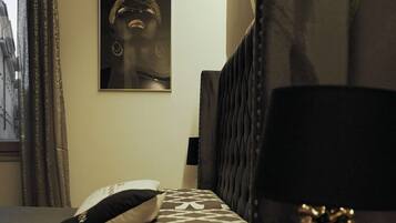 Una camera d'albergo con un letto, un comodino con una lampada, un cuscino, un cuscino a fantasia, un quadro appeso al muro e una tenda.