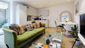A modern living room with a green sofa, a glass coffee table, and a kitchen in the background. - Boho-Chic Escape, by Veeve (London)