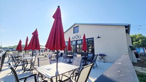 Un espace extérieur aménagé avec des tables et des parasols rouges, un bâtiment blanc portant l’inscription « AFOG », et un ciel dégagé.