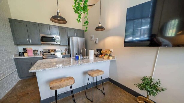 Une cuisine moderne avec des meubles gris, un grand îlot central doté de tabourets, un téléviseur à écran plat et des plantes suspendues.