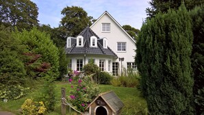 Una casa de dos pisos con un techo pronunciado, rodeada de abundante vegetación y una caseta para perros en el jardín delantero.
