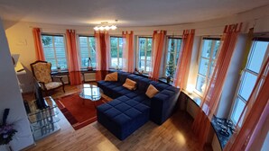 Un salón con un sofá seccional azul oscuro, una mesa de centro de vidrio y una alfombra roja. Hay varias ventanas con cortinas naranjas, un radiador y una lámpara sobre una mesa.